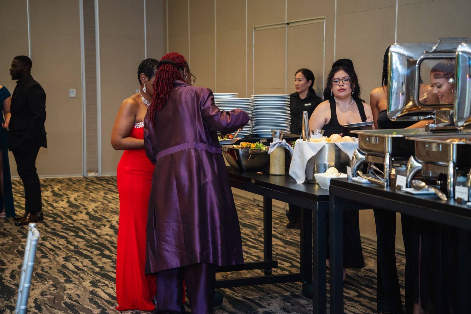 People in formal attire gather at a conference or event venue with tables and carpeted flooring.