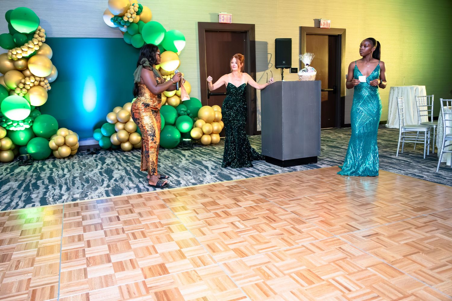 Elegant ballroom setup with green balloon decorations and gold accents illuminated by colorful spotlights.