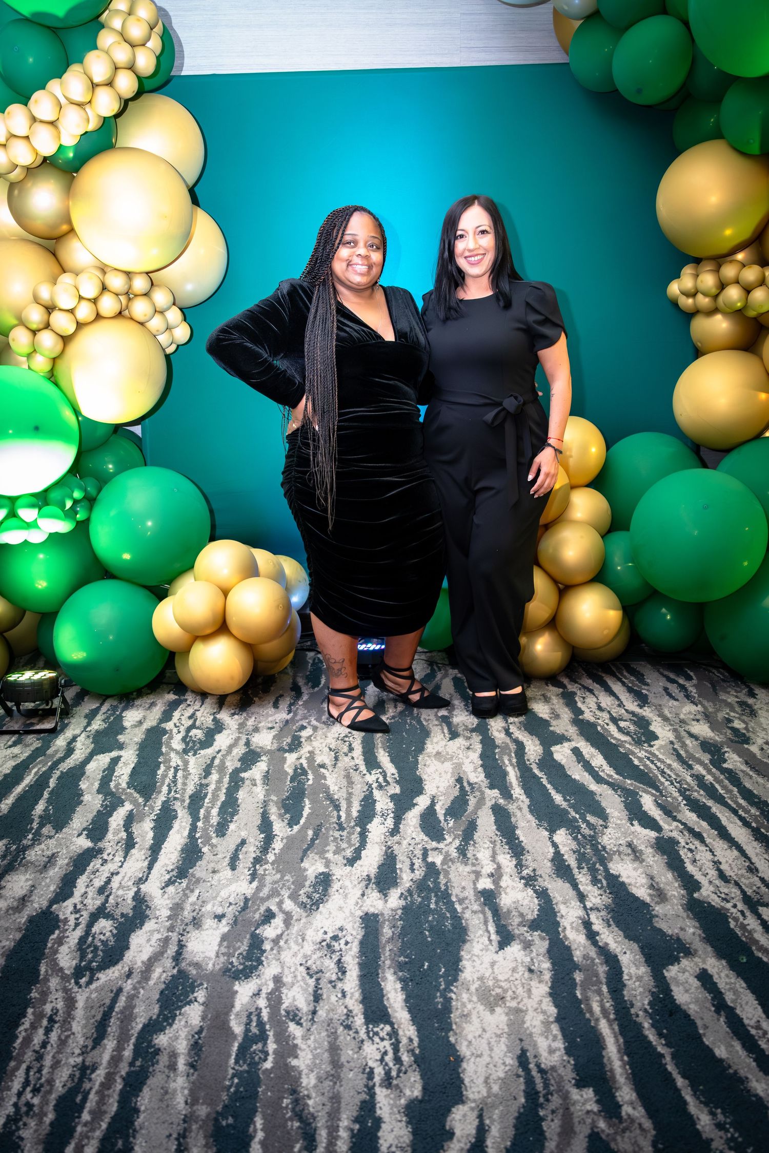 People posing against a festive backdrop with green and gold balloon decorations and a textured gray floor.
