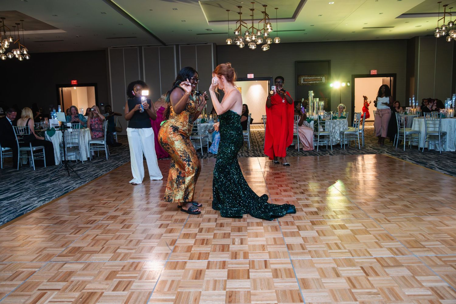 People dancing and socializing in a spacious ballroom with hardwood floors and ambient lighting.