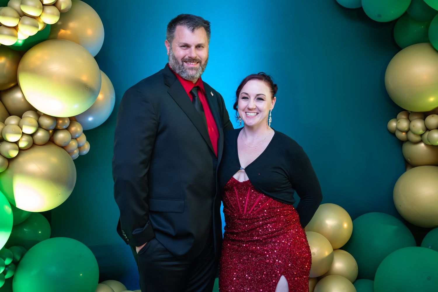 A couple in formal attire poses against a festive backdrop with green and gold bokeh lights and ornaments.