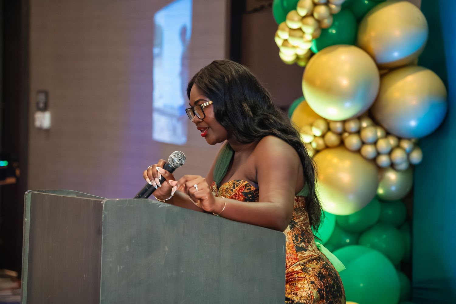 Speaker in a gold sequined dress stands at podium with green and gold balloon arch decoration in background.