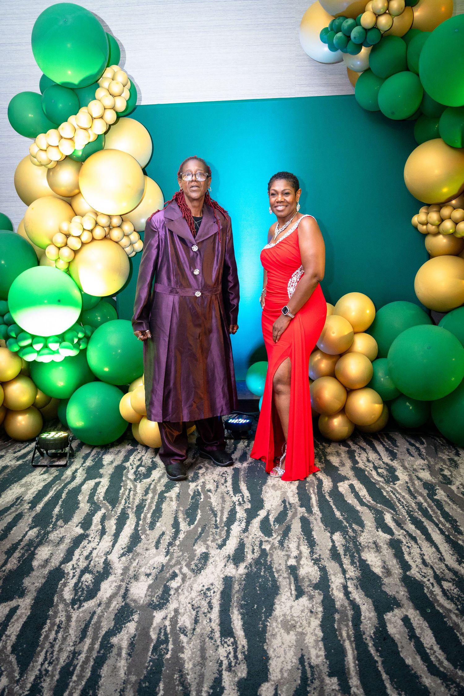 A couple poses in front of a festive balloon arch with green and gold decorations at a celebration event.