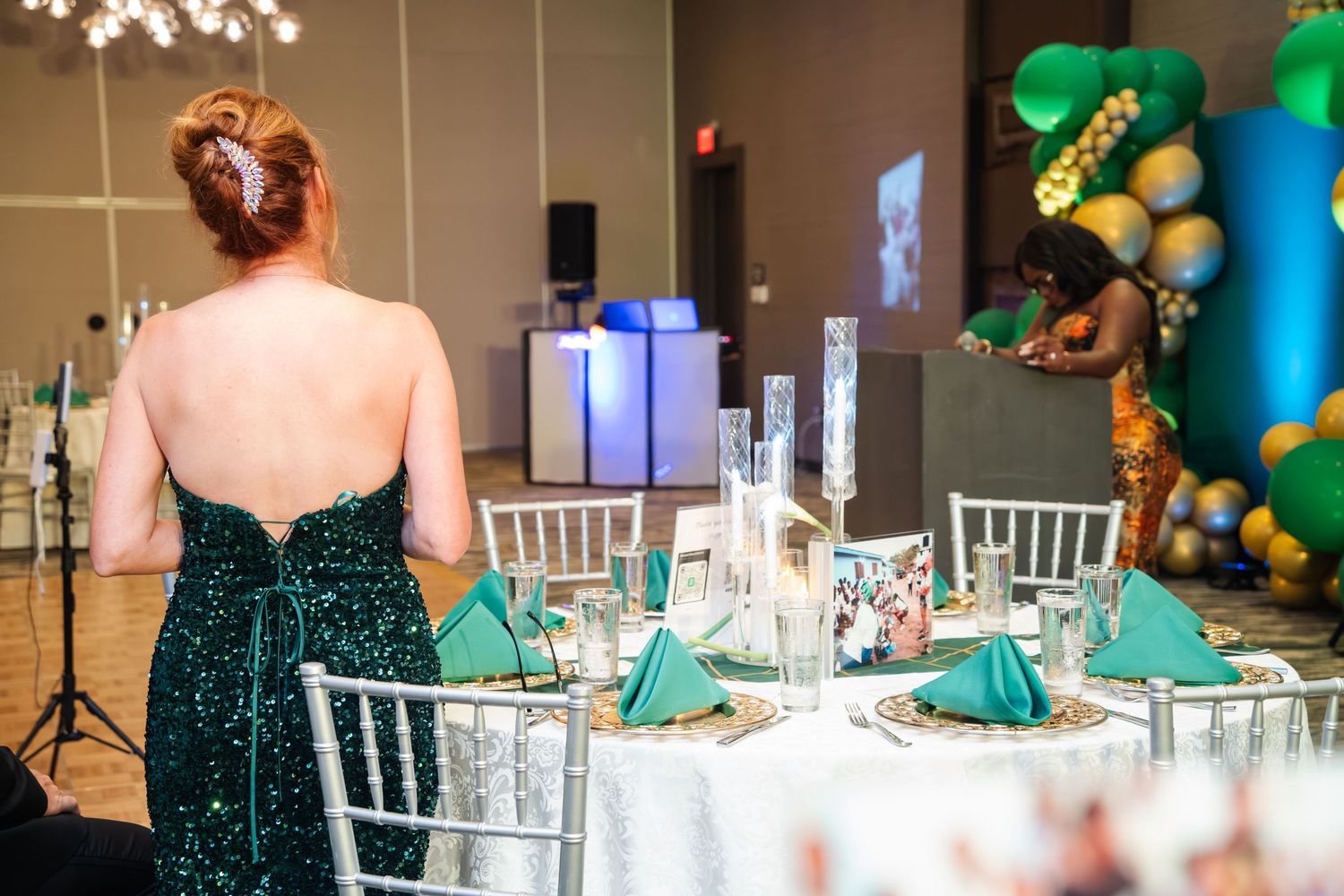 Elegant wedding reception setup with emerald green decor, white chairs and balloon arch in a banquet hall.