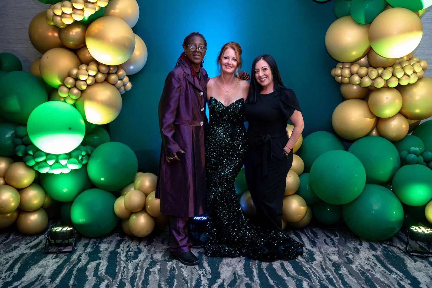 Three people pose together in front of a decorative green and gold balloon arch at an elegant event.