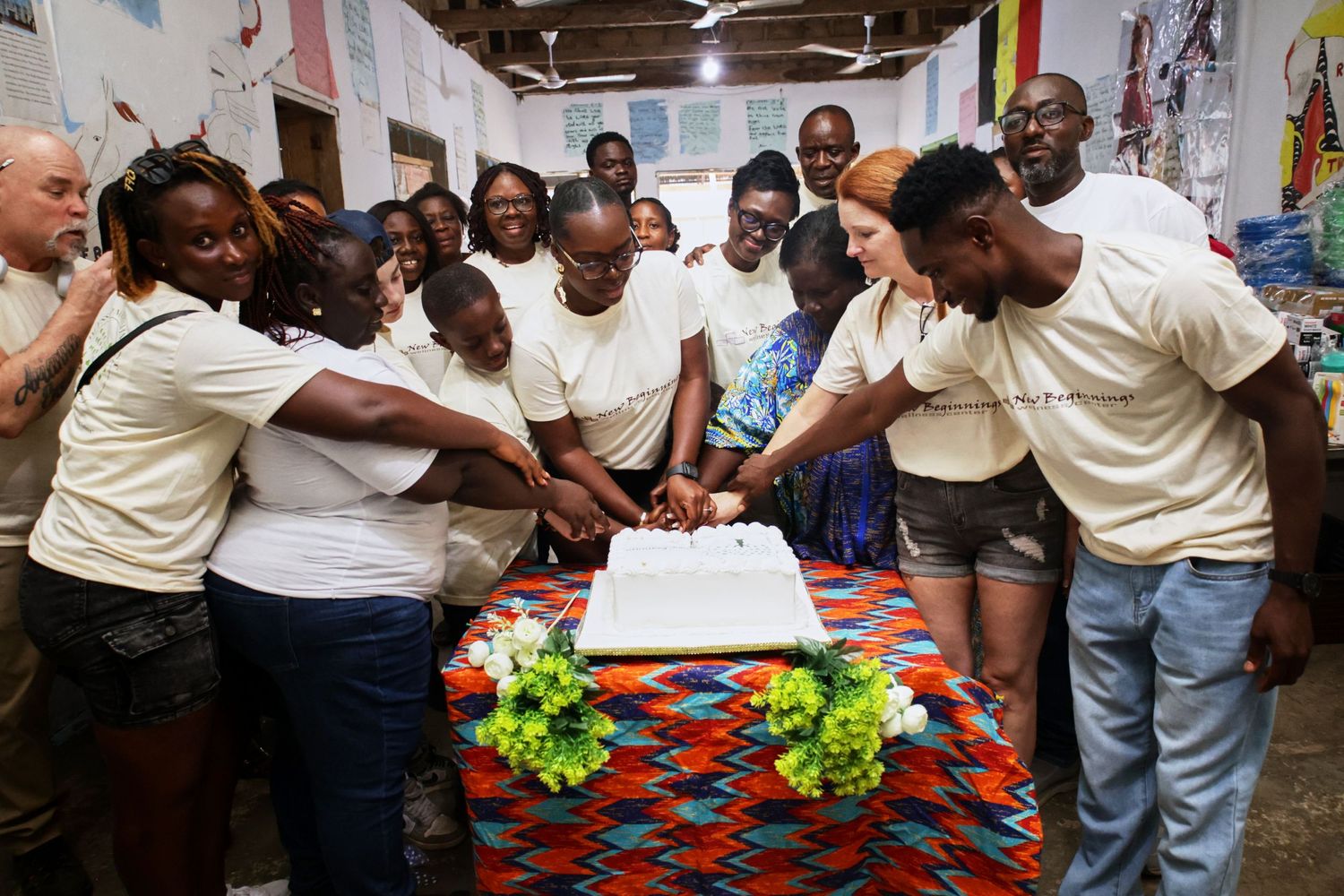Celebration scene with people surrounding a decorated cake table with colorful tablecloth.