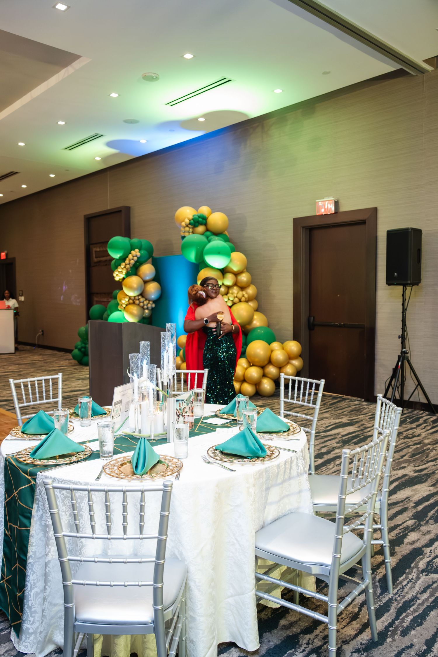 Elegant banquet hall setup with white Chiavari chairs, round tables, and gold balloon decorations against blue-lit walls.