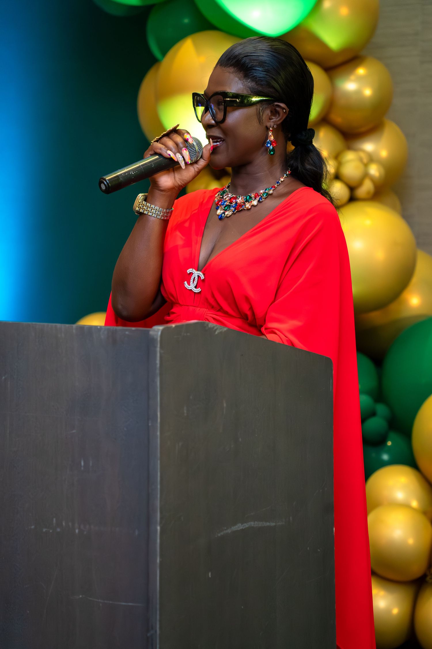 Person in red delivers a speech at a podium with decorative gold and green balloon arrangements behind them.