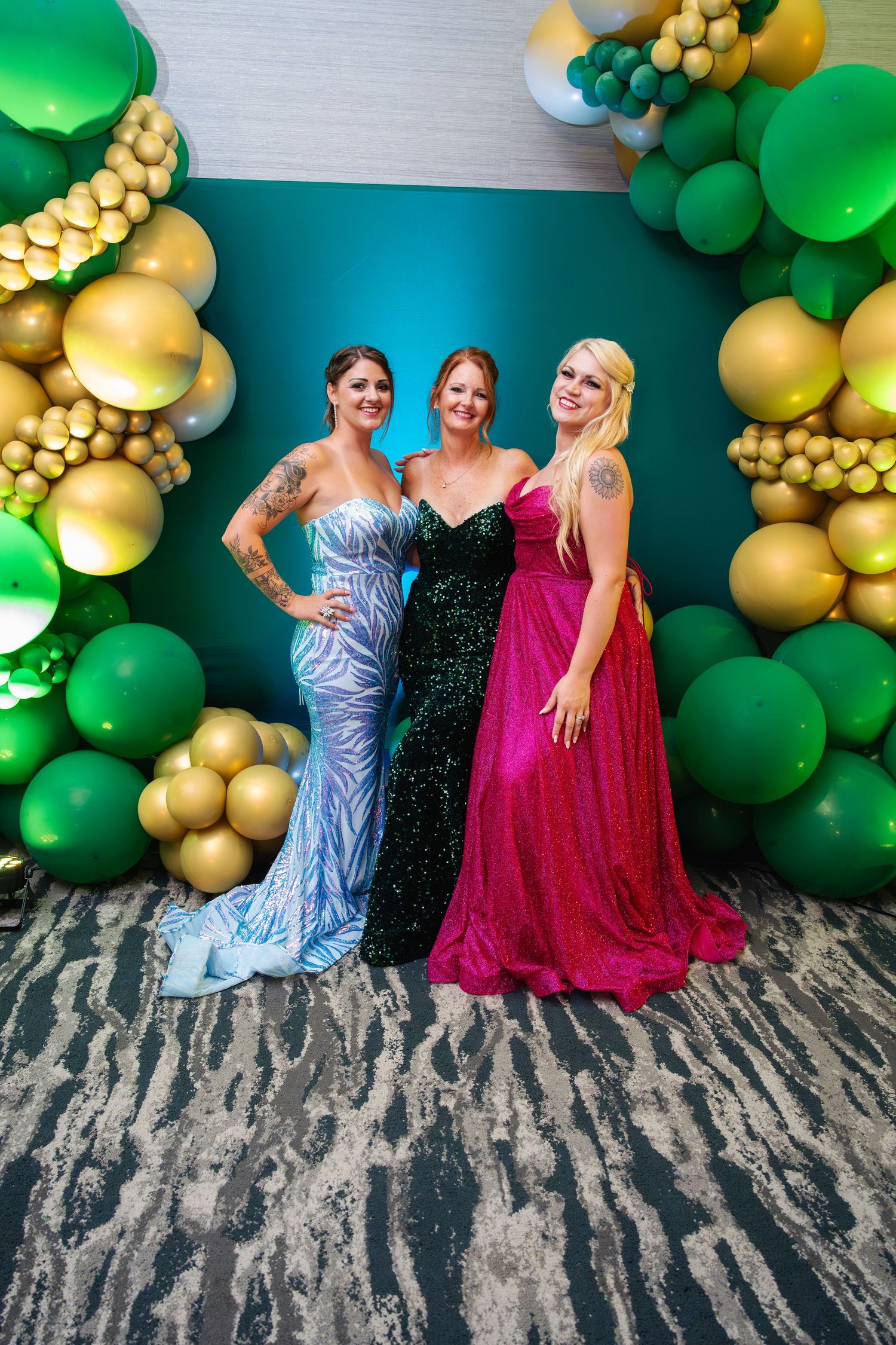 A festive party scene with green and gold balloon arch decorations against a wall with three people posing together.