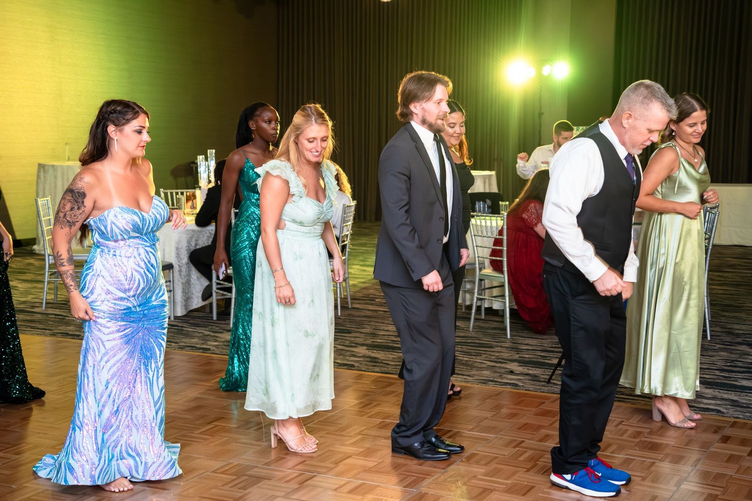 Wedding guests dance together on a wooden floor at an indoor reception venue with yellow lighting.