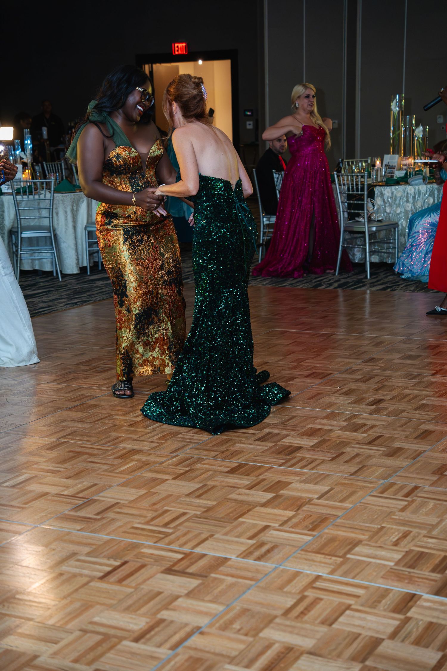 Guests in elegant evening gowns dance on a parquet floor during a formal event in a ballroom.