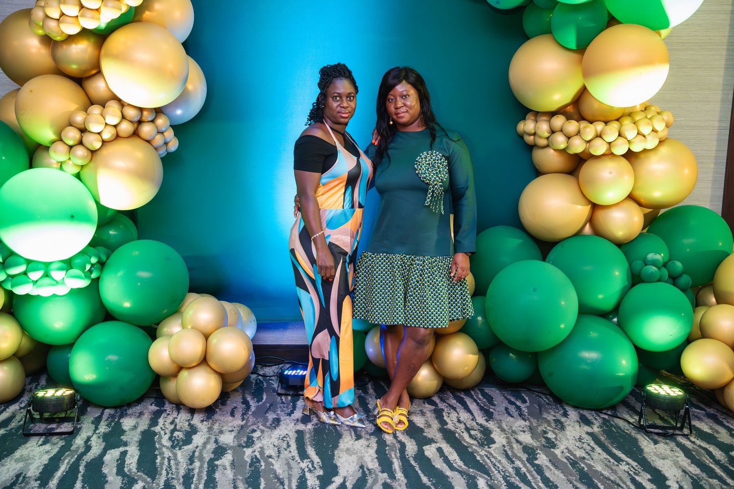 Two people pose in front of a decorative balloon arch with green and gold balloons against a blue backdrop.