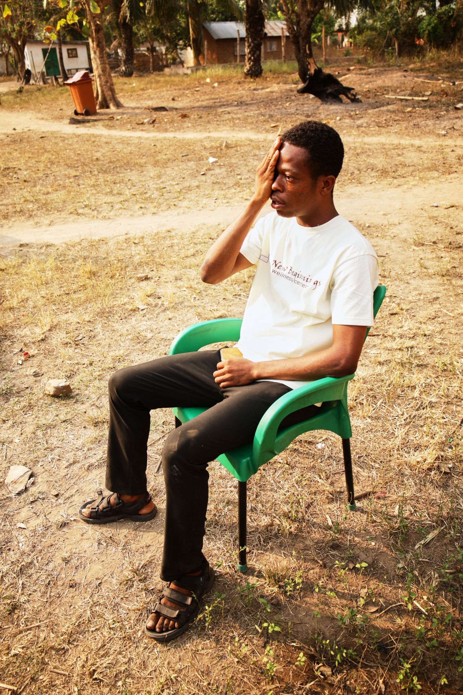 Person in white t-shirt sitting thoughtfully in green plastic chair outdoors.