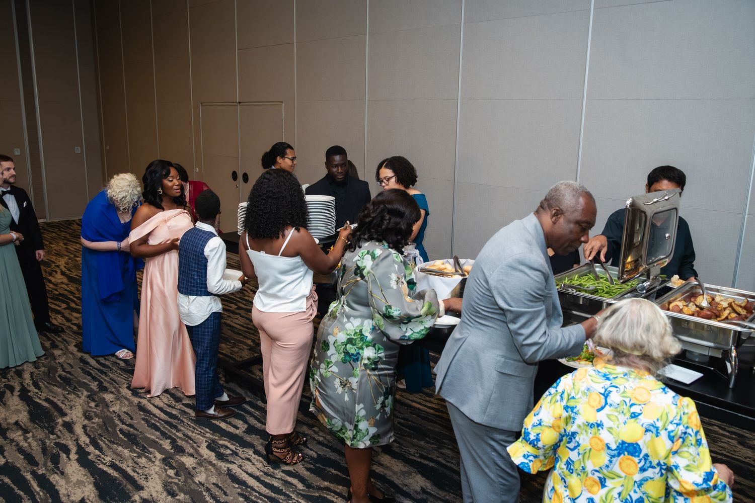 People gather around food tables at an indoor social event with gray walls and carpeting.