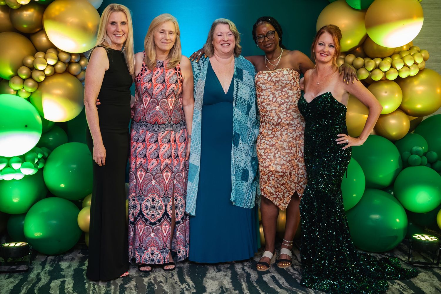 Group poses in front of a festive balloon wall with green and gold decorations at a celebration event.