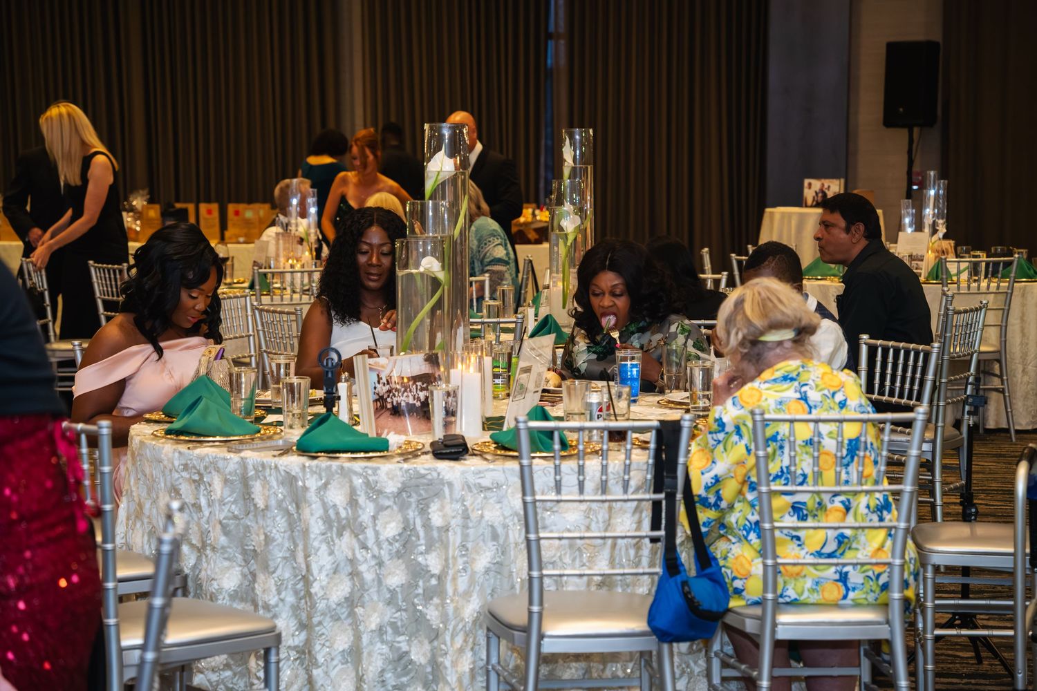 Wedding guests sit at decorated tables during a reception in a large banquet hall.