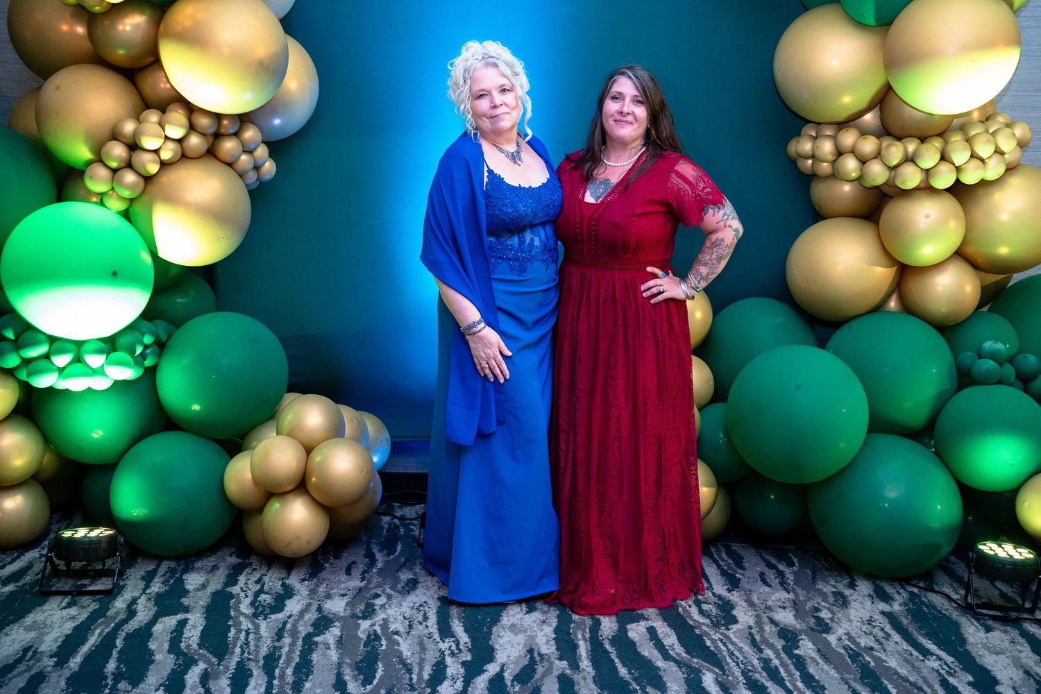 Two people in elegant long dresses pose in front of a festive balloon arch with gold and green decorations.