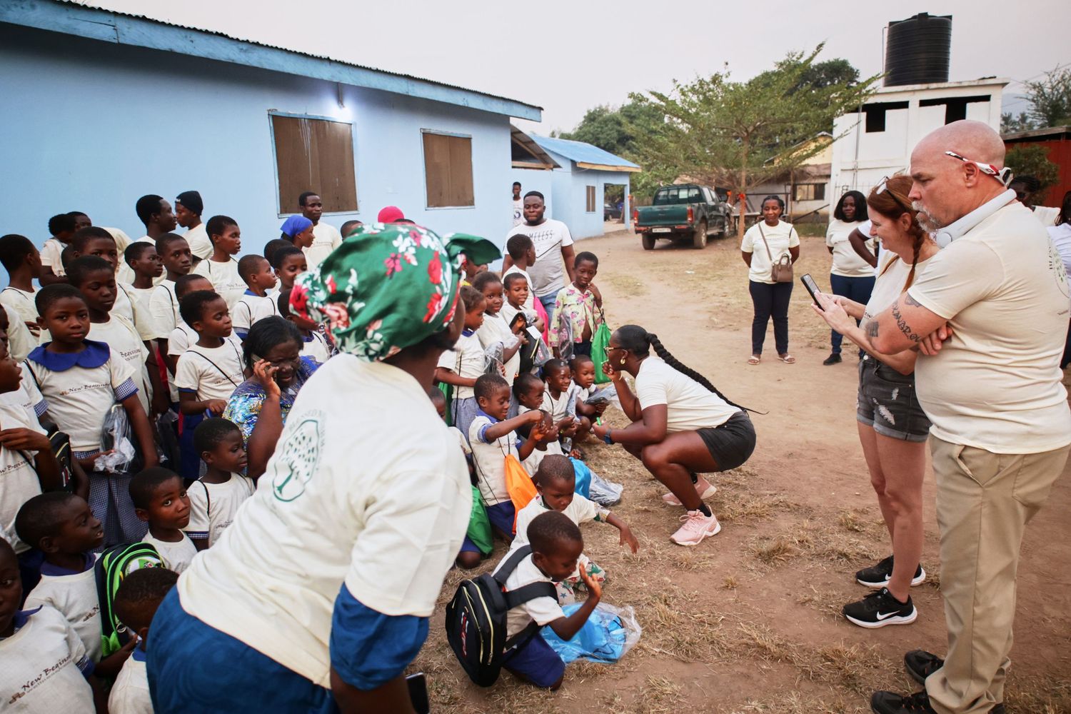Outdoor gathering of students receiving supplies while volunteers document the event.