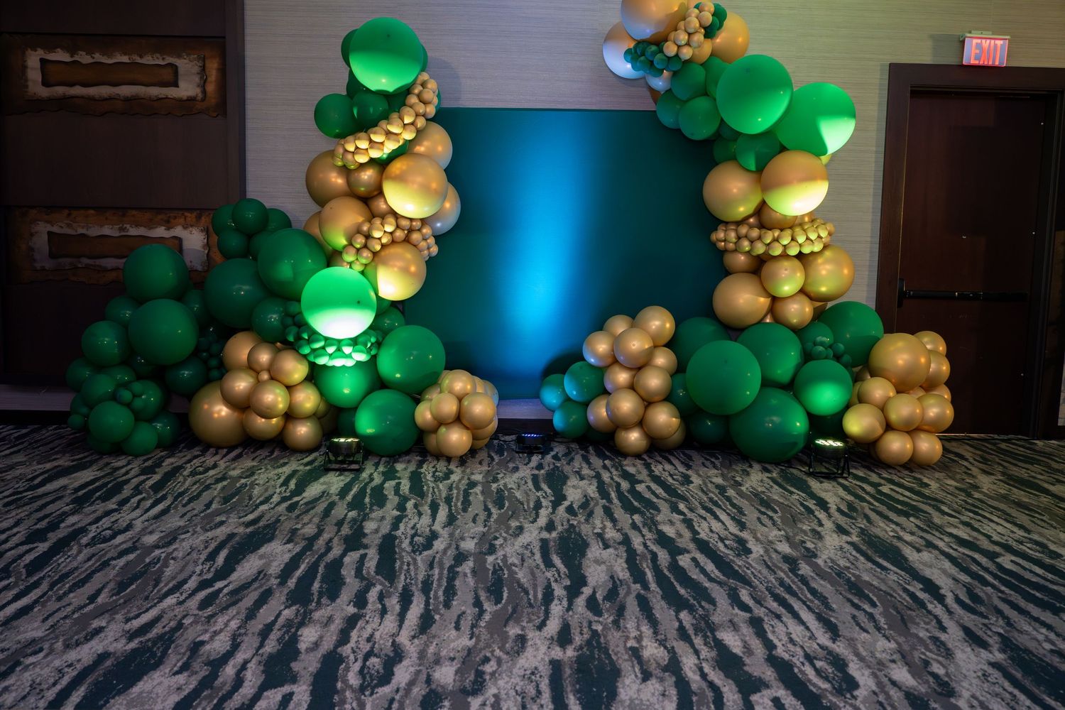Green and gold balloon arch decorations frame a blue backdrop against a patterned carpet floor.