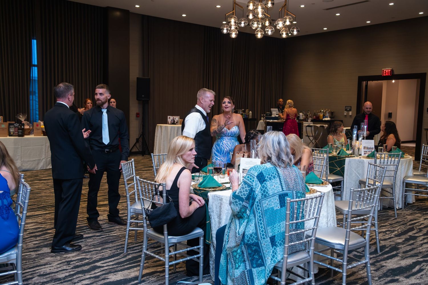 Wedding reception guests gather around elegant silver chiavari chairs and tables under a crystal chandelier in a modern ballroom.