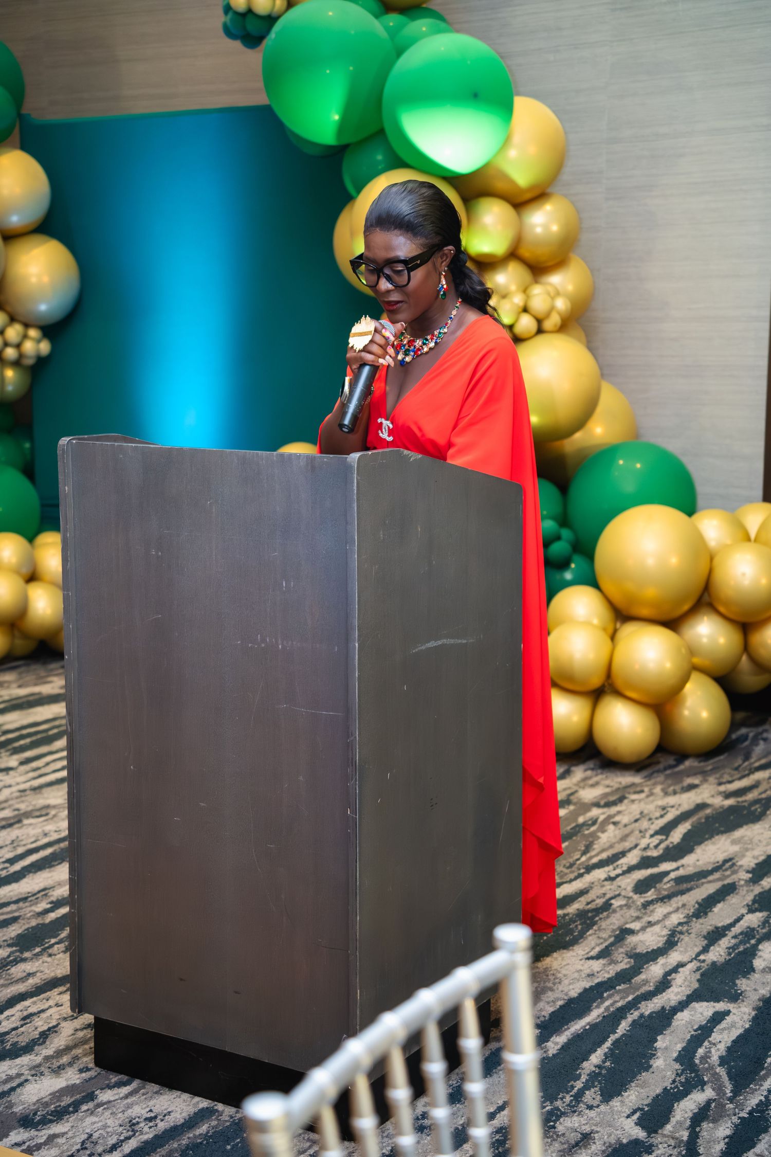 A person in a red top sits at a dark podium against a backdrop of green and gold balloon decorations.