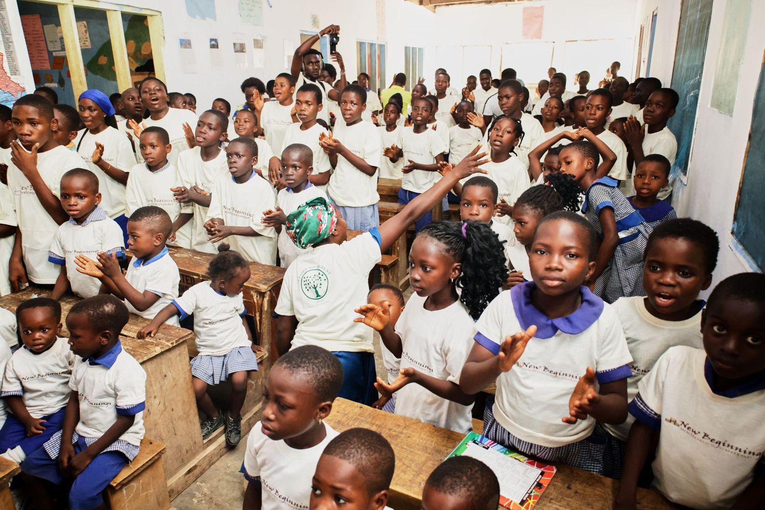 Large group of enthusiastic students in white t-shirts gathered in a classroom raising their hands excitedly.