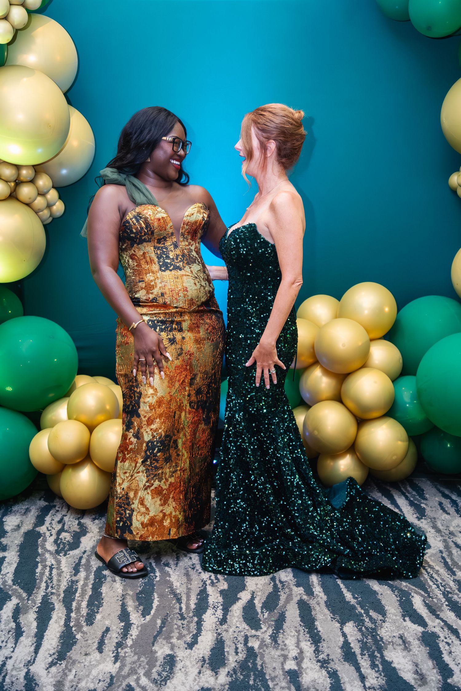 Two people in formal dresses pose against a teal wall decorated with gold and green balloon garlands.