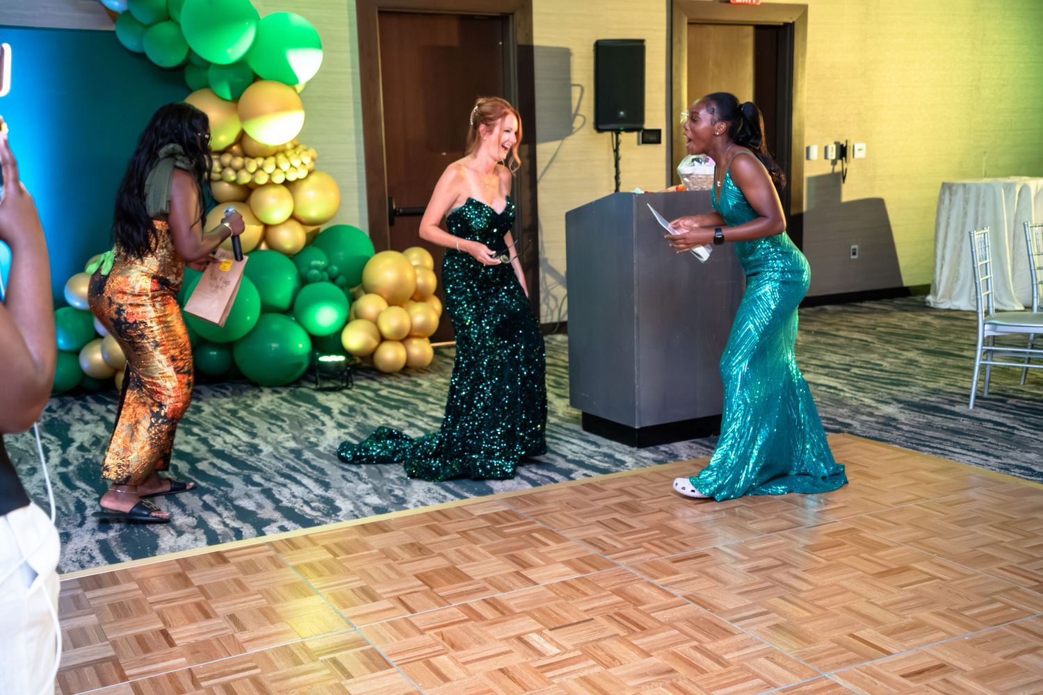 People in sparkly evening gowns dance on a wooden floor with green and gold balloon decorations in the background.
