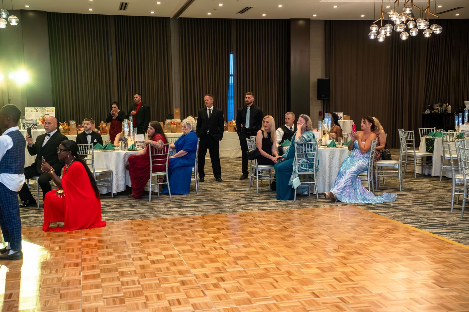 Panoramic view of a formal ballroom event with a checkered dance floor and elegant chandeliers overhead.