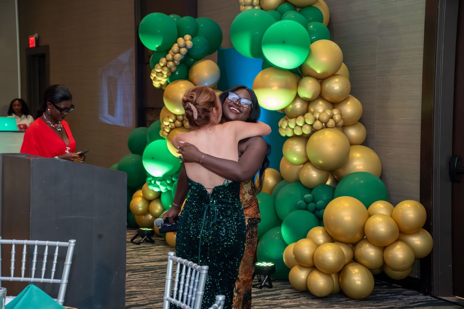 A couple embraces in front of an elegant balloon arch display featuring green and gold balloons at an event venue.
