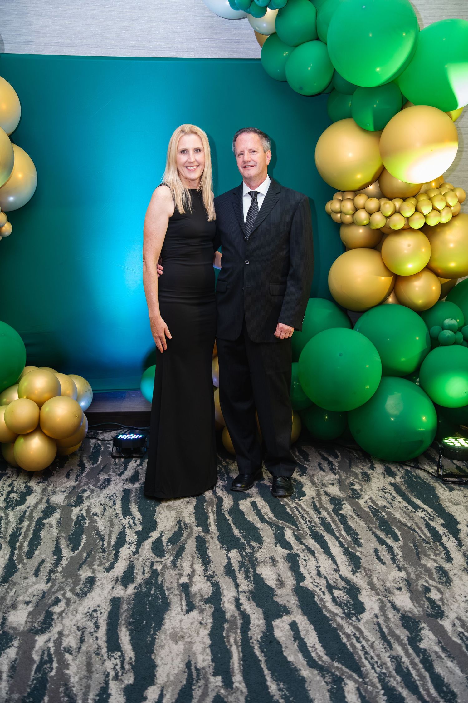 Couple stands in front of green and gold balloon arch decorations with teal backdrop at formal event.