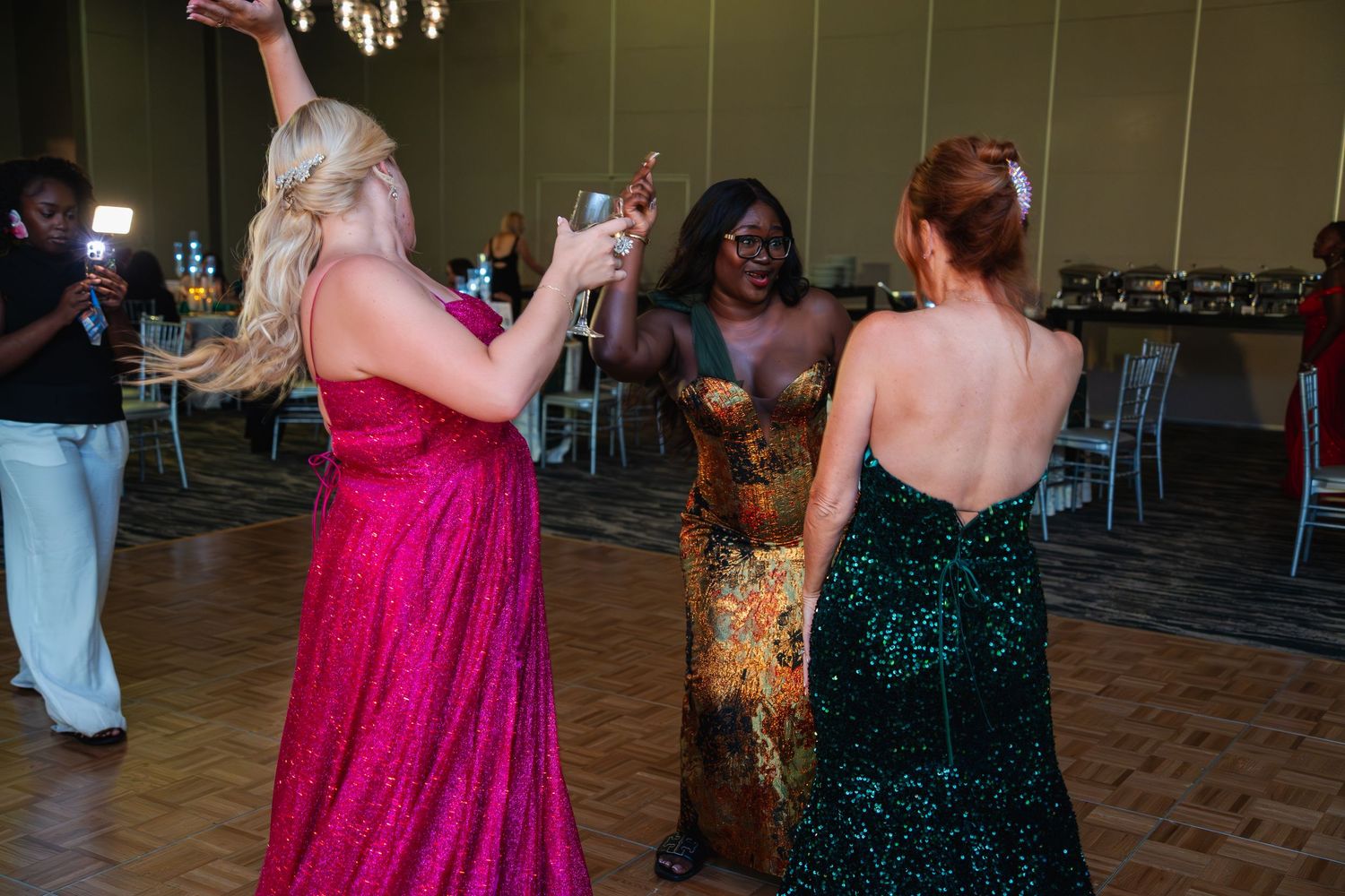 Three women in formal evening gowns dance together on a parquet floor at an elegant event.
