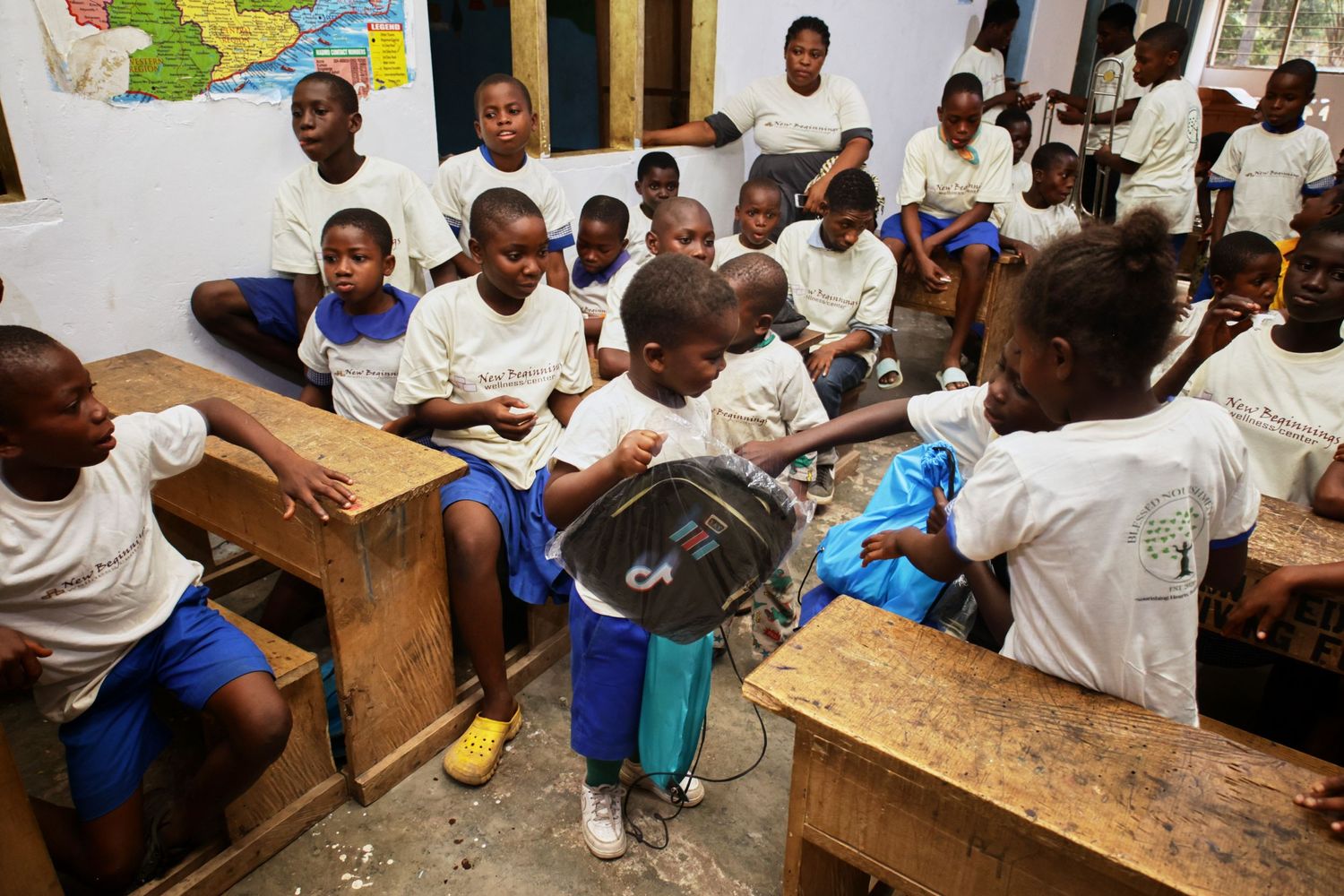 Students in white uniforms and blue shorts sit at wooden desks in a classroom with a wall map.