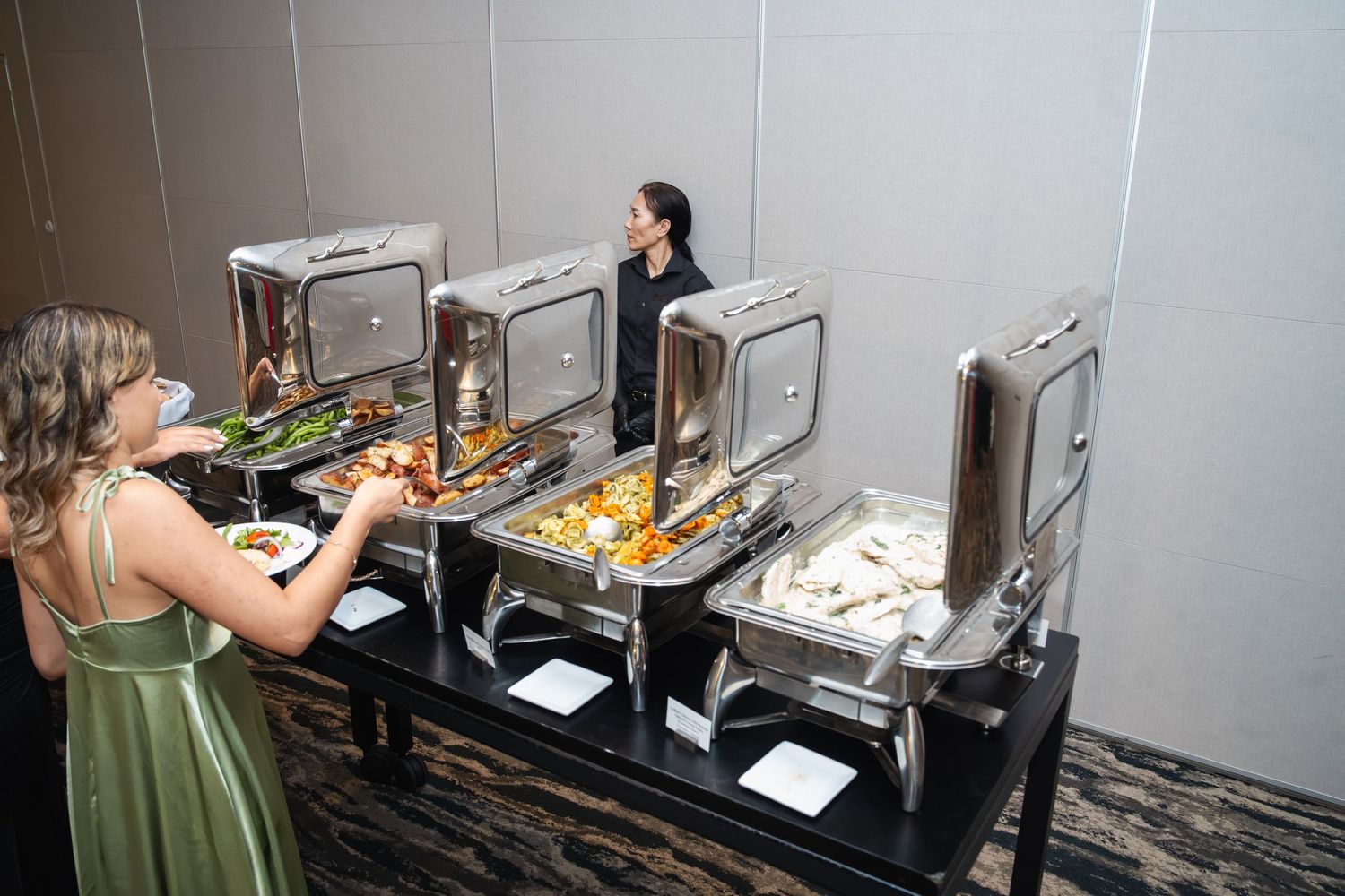Two people prepare food at stainless steel buffet stations in a commercial kitchen setting.