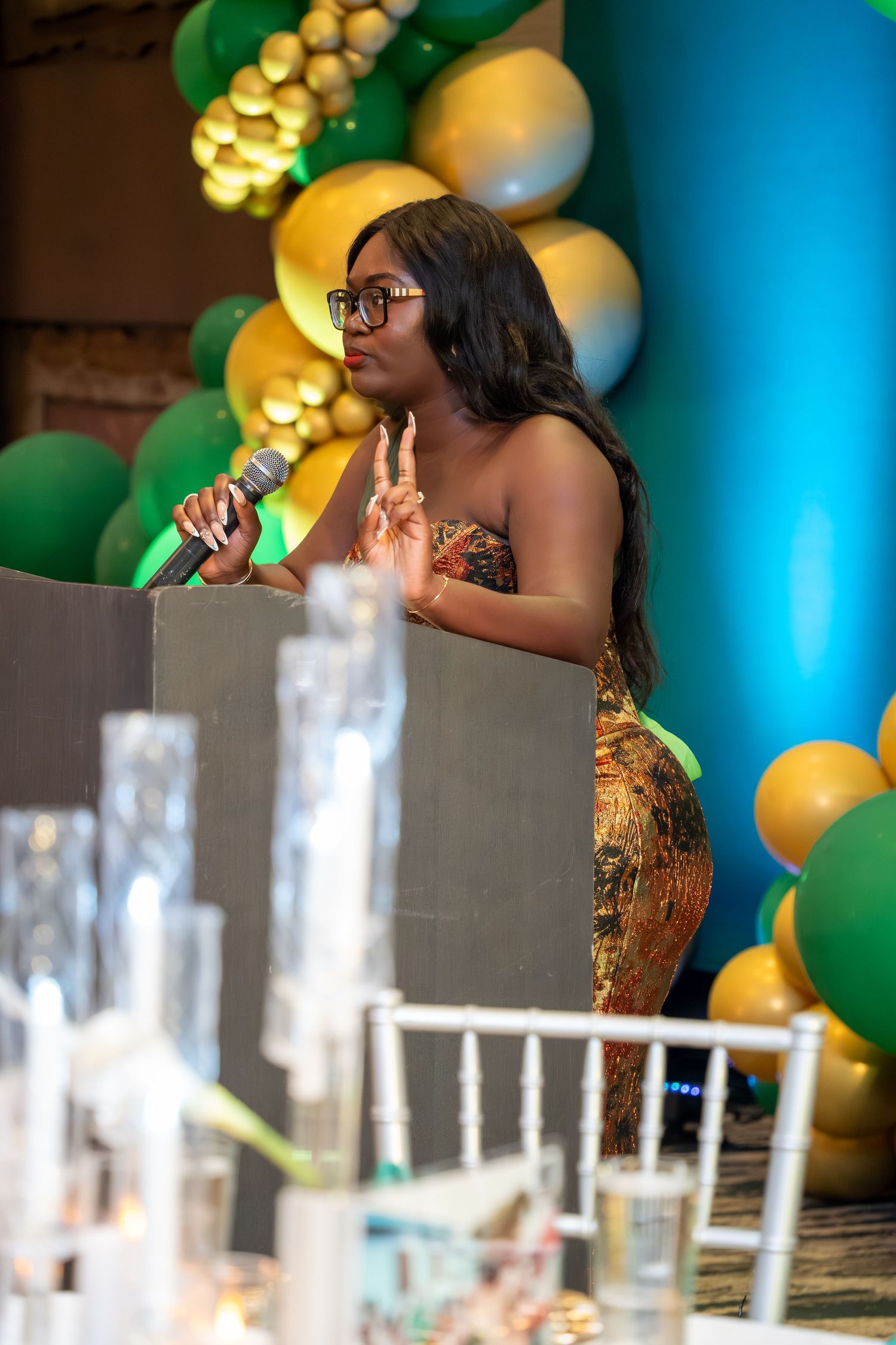 A speaker in a formal dress addresses guests at a decorated event venue with balloons and table settings.