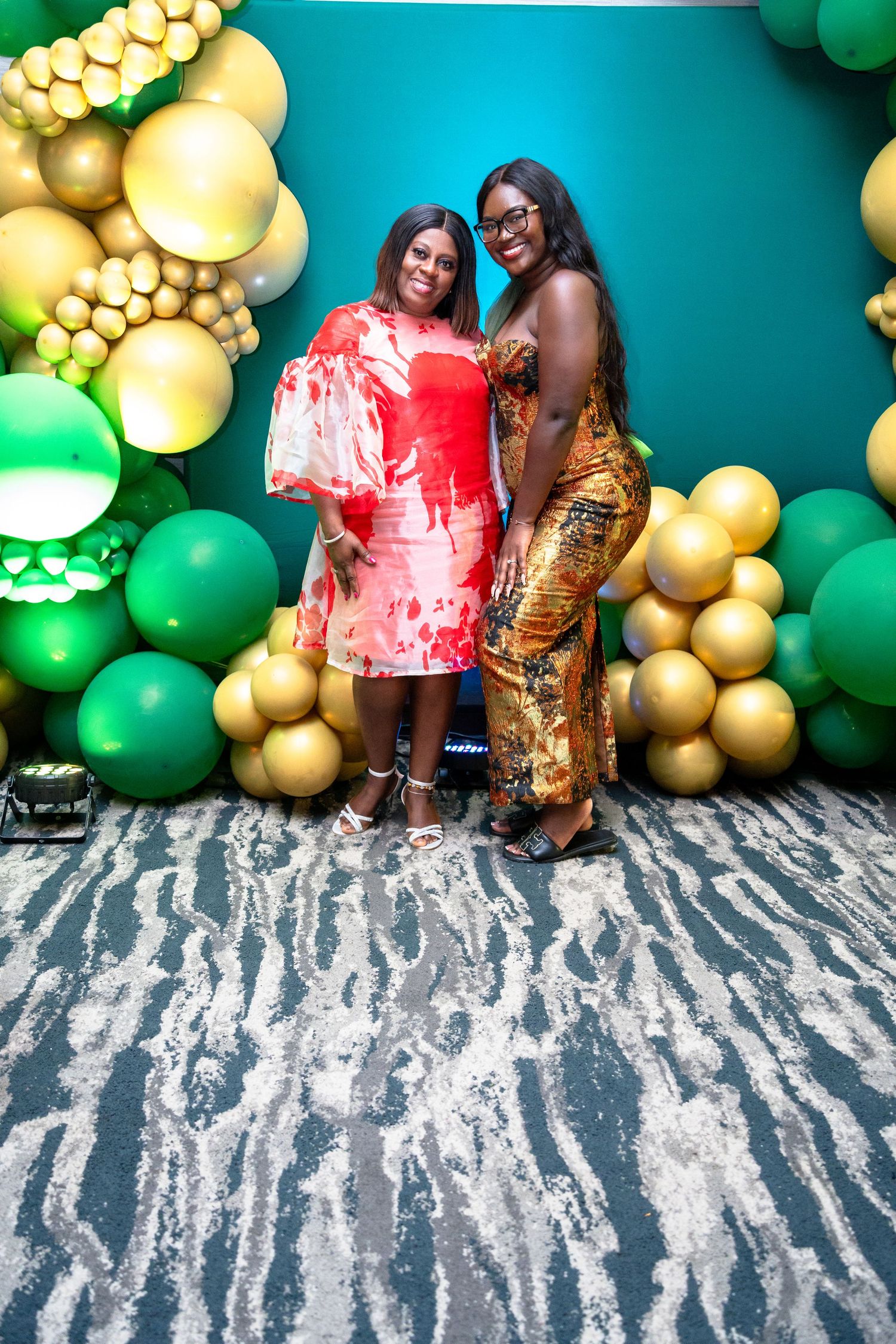 Two people pose against a festive wall decorated with green and gold balloon garlands and a zebra print floor.