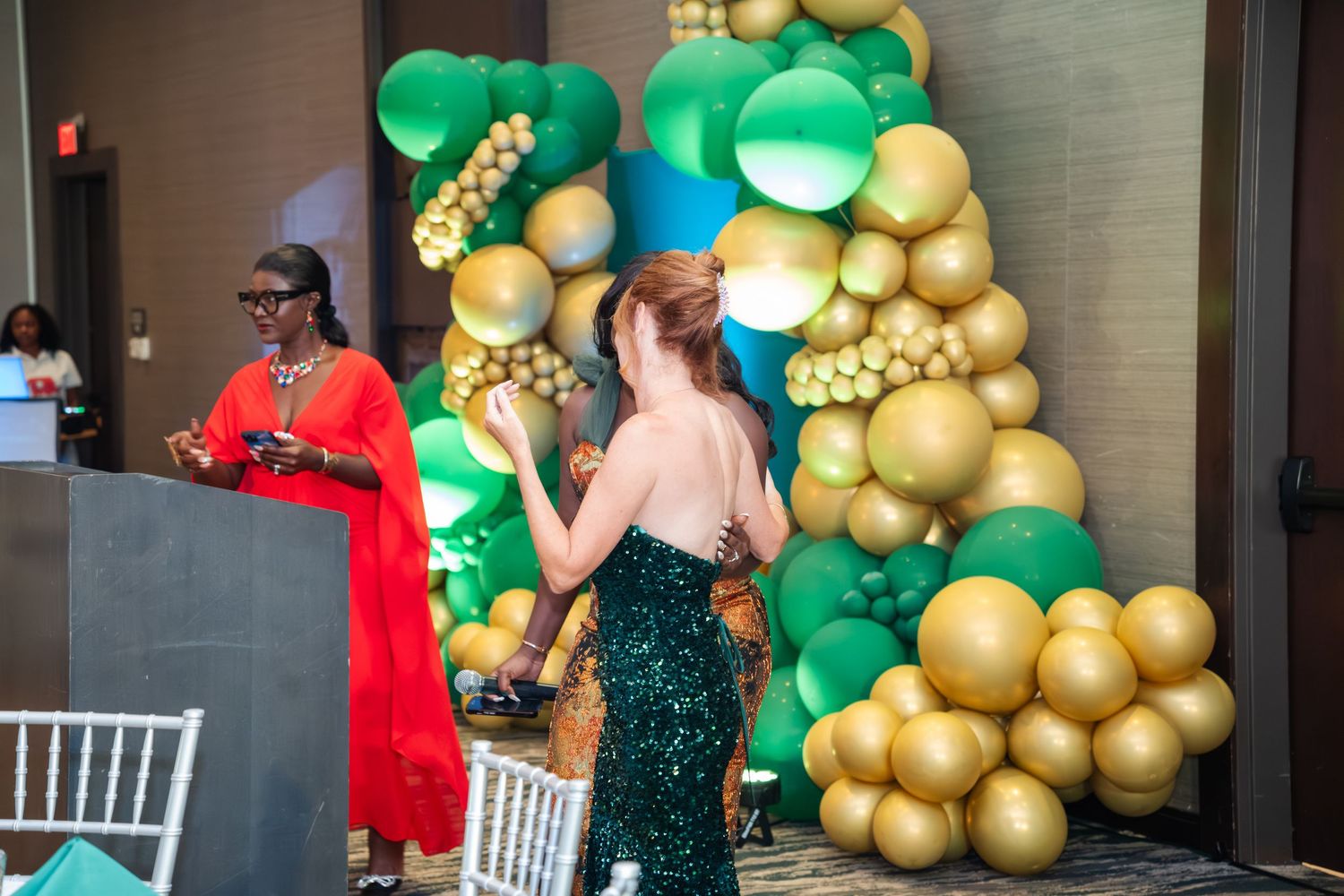A festive party setup with gold and green balloon decorations against a wall with people mingling in formal attire.