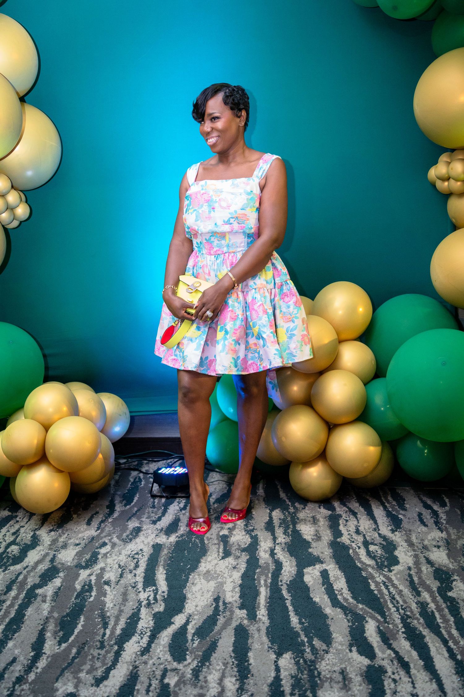 A person in a floral dress poses against a backdrop of gold and green balloons with a textured wall.