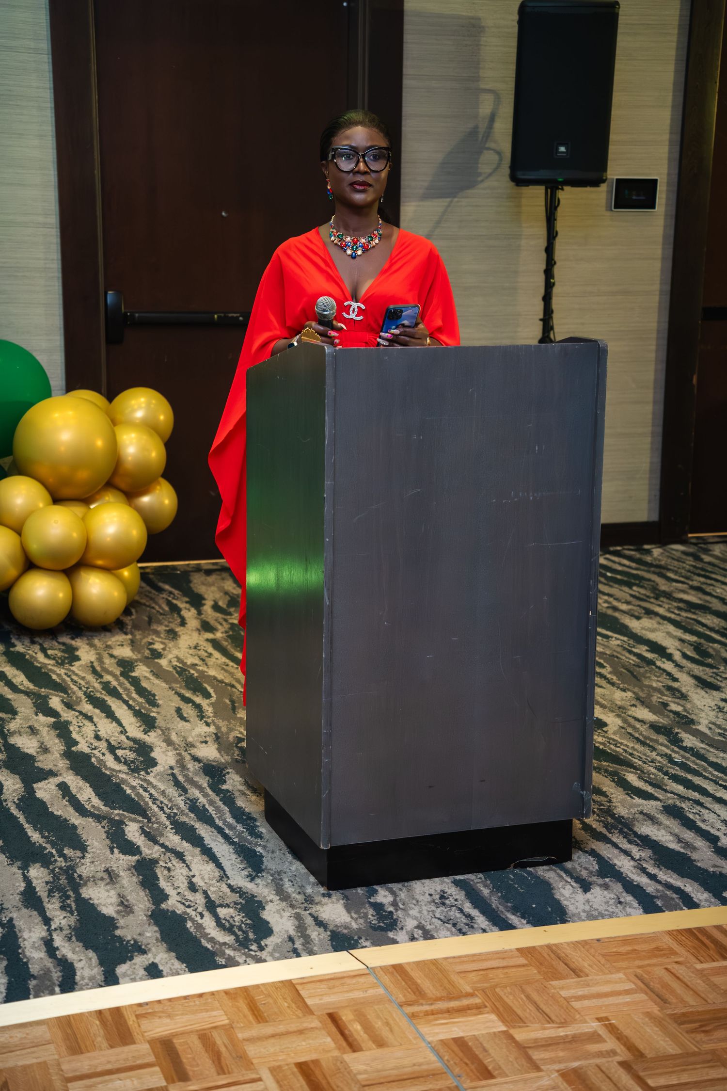 Black cube-shaped podium sits on patterned carpet with gold balloons in background and red decor accents.