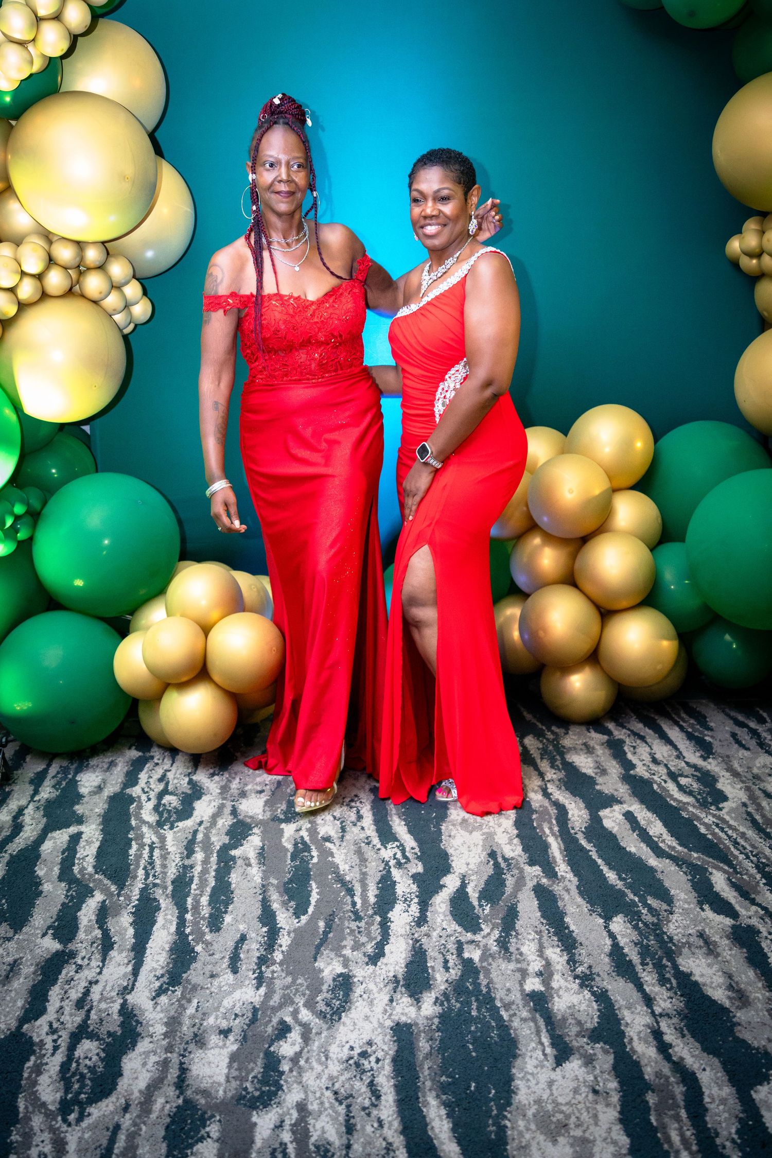 Two women in matching red formal dresses pose together against a backdrop of green and gold balloon decorations.