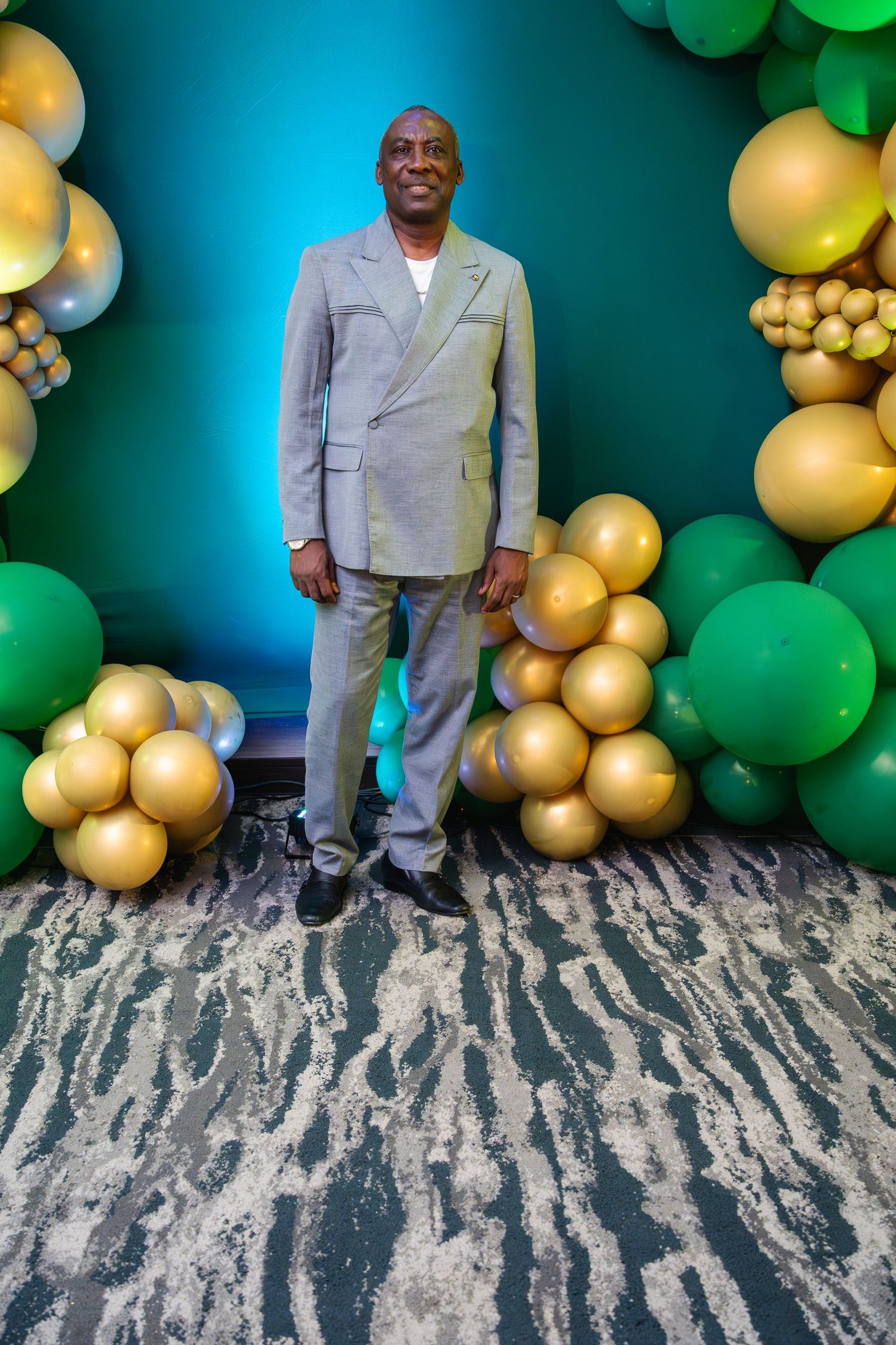 A person in a gray suit poses against a backdrop decorated with green and gold balloon arrangements.