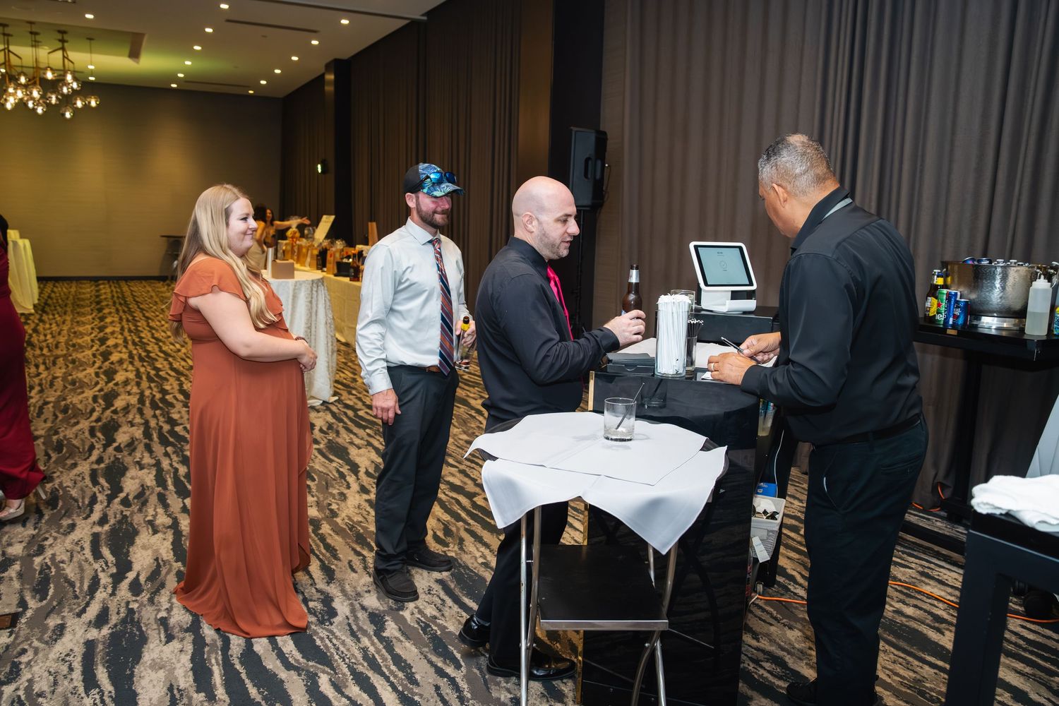 Event attendees interact with staff at a cocktail reception in an elegant hotel ballroom with patterned carpet.