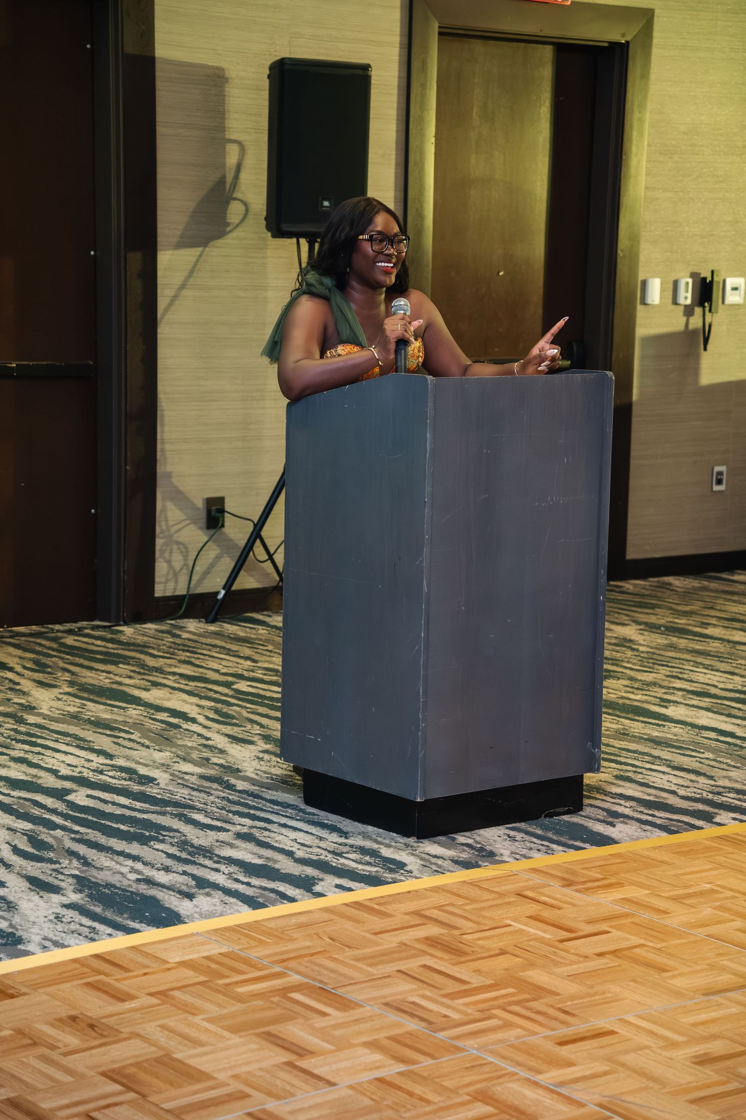A person stands behind a dark gray podium on a patterned carpet floor in a conference room setting.