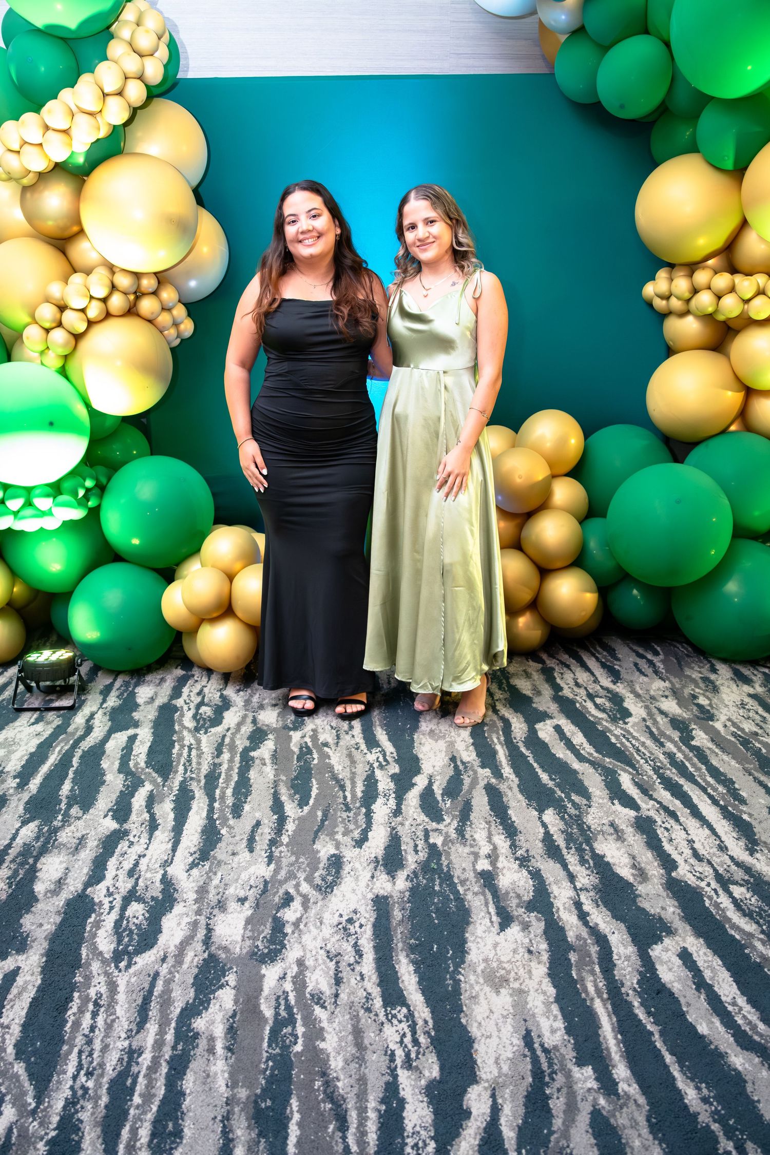 A festive photo backdrop with green and gold balloon garland against a teal wall and zebra-striped floor.