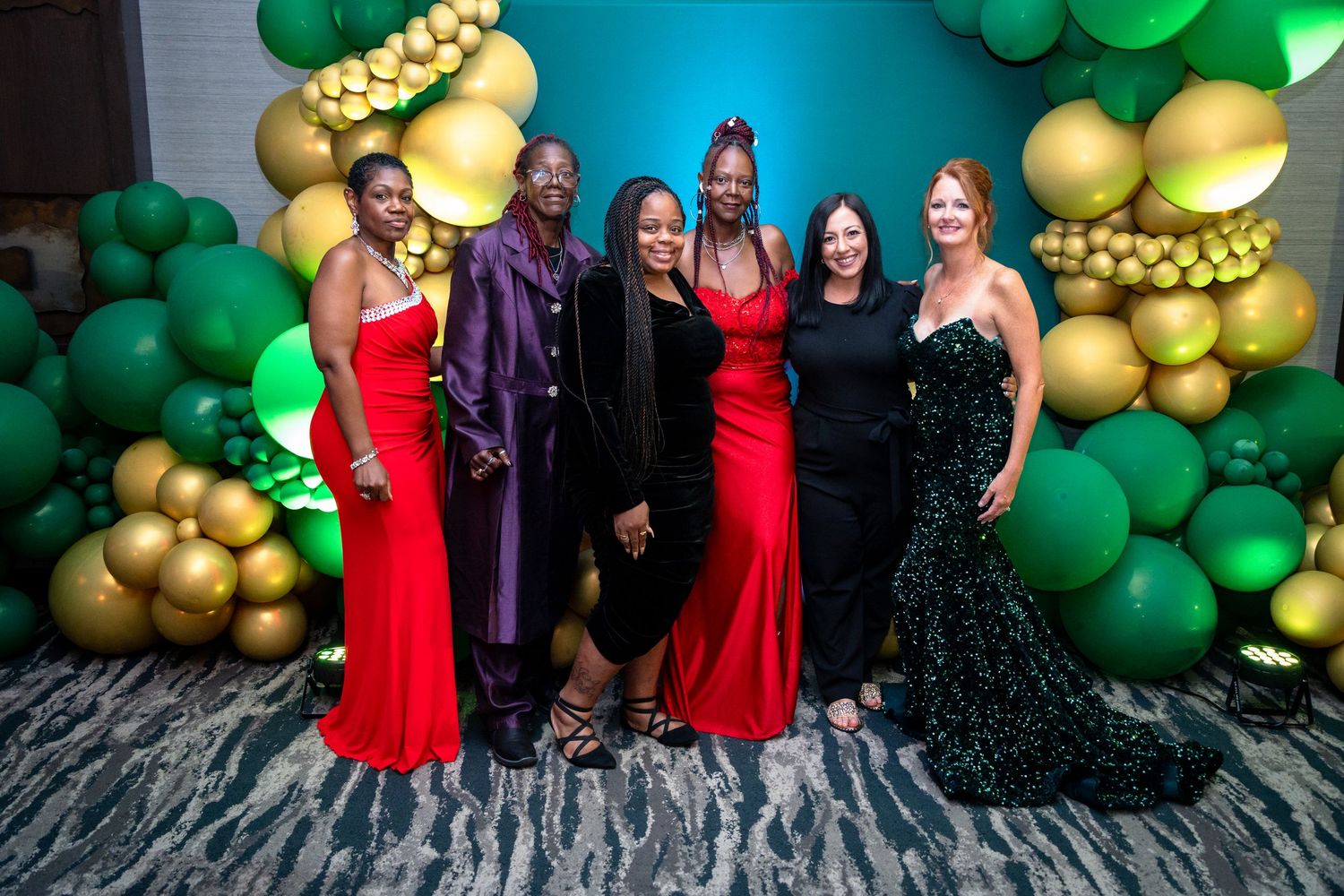 A group poses in formal evening gowns against a decorative backdrop of green and gold balloons.