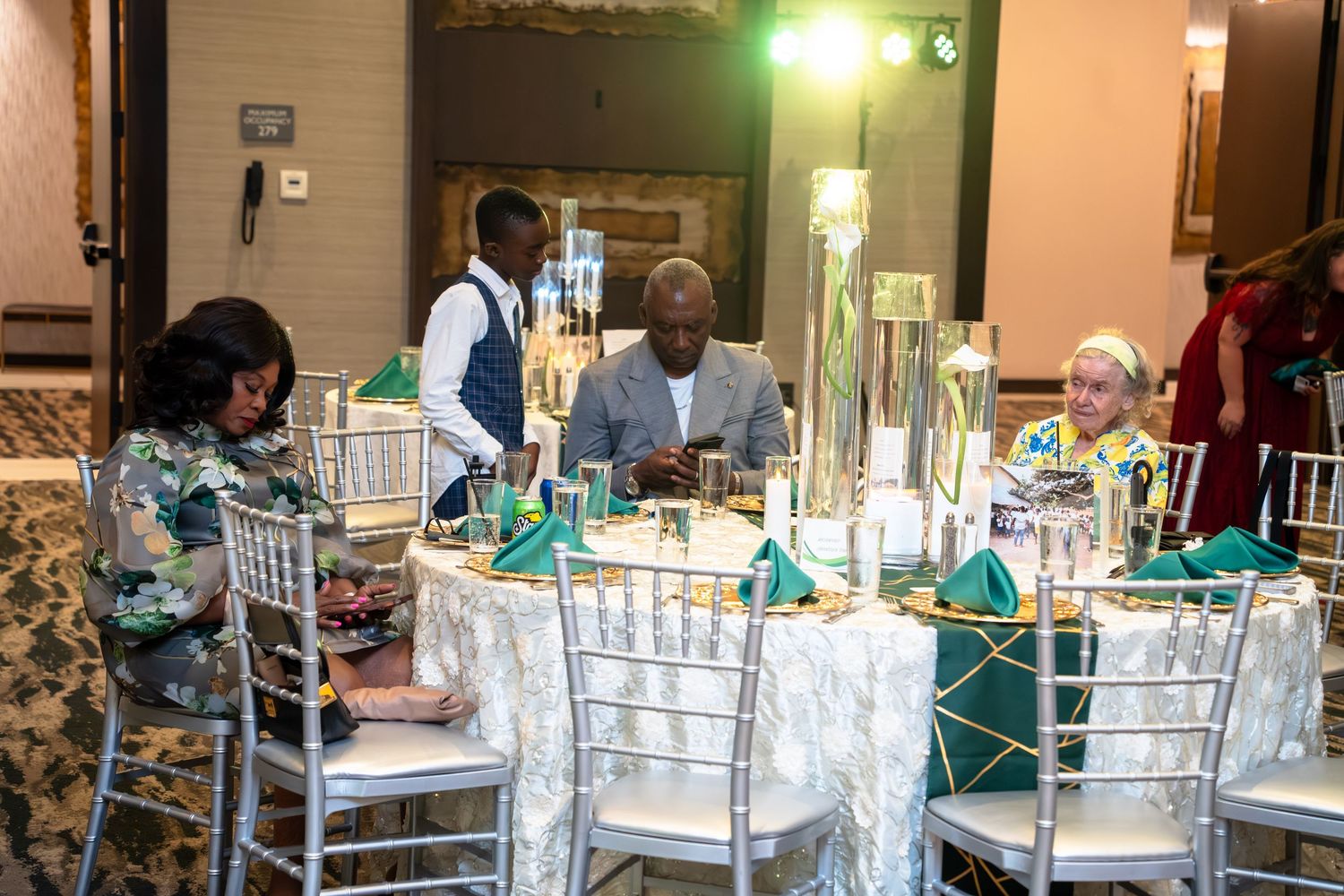 A formal event venue with white Chiavari chairs and elegant table settings illuminated by soft green lighting.