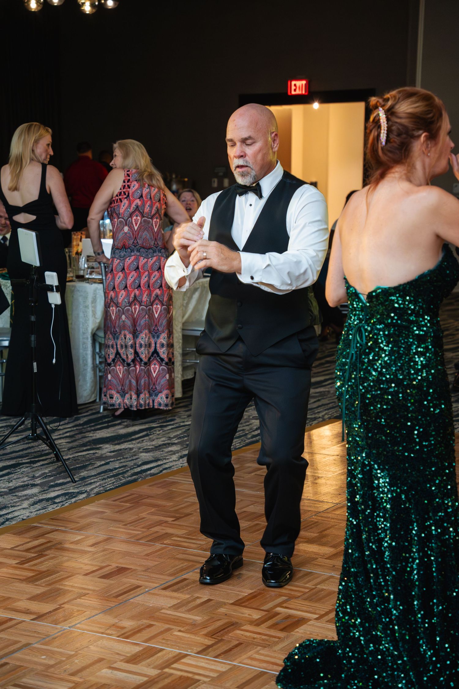 Dancers in formal attire move across a parquet dance floor at an elegant event with a green sequined dress visible.