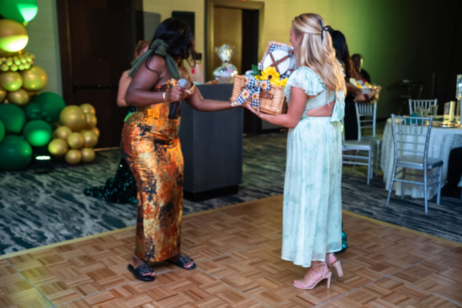 Dancers move on a wooden dance floor at an indoor event with green lighting and gold balloon decorations.