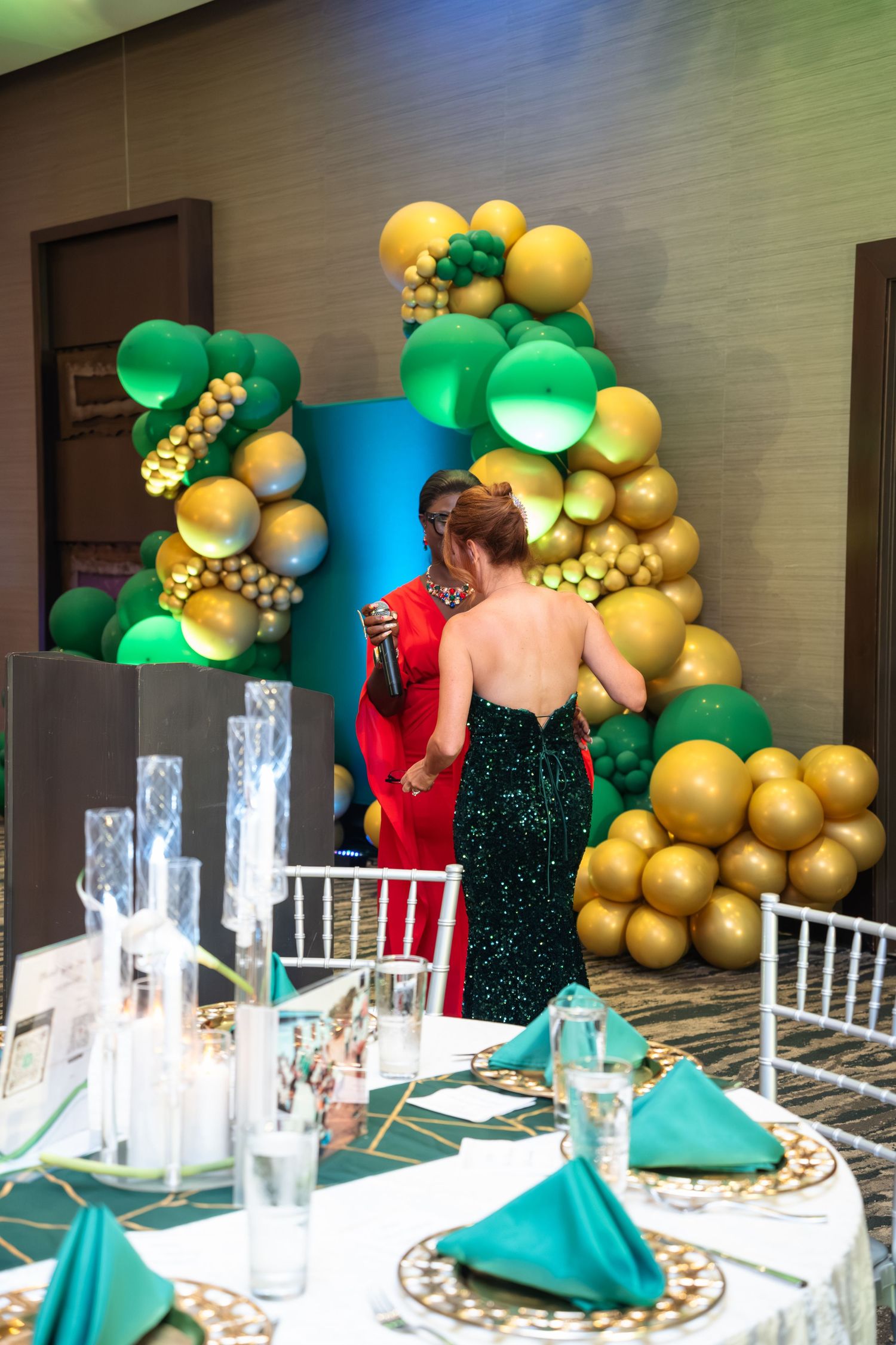 Elegant event setup with teal napkins and gold balloon arch featuring green accents against textured wall.