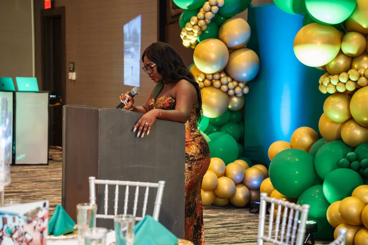 A festive event space decorated with green and gold balloon garlands and white chairs for a celebration.